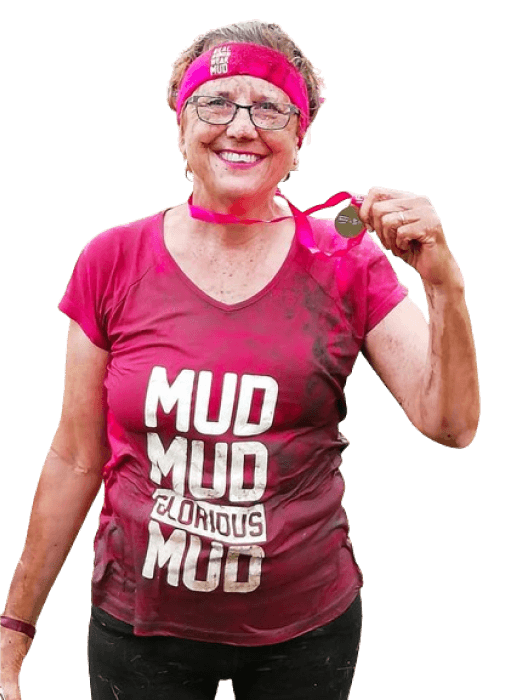 Karen, smiling and covered in mud, holding her medal after completing Pretty Muddy, raising money for Breast Cancer Research UK